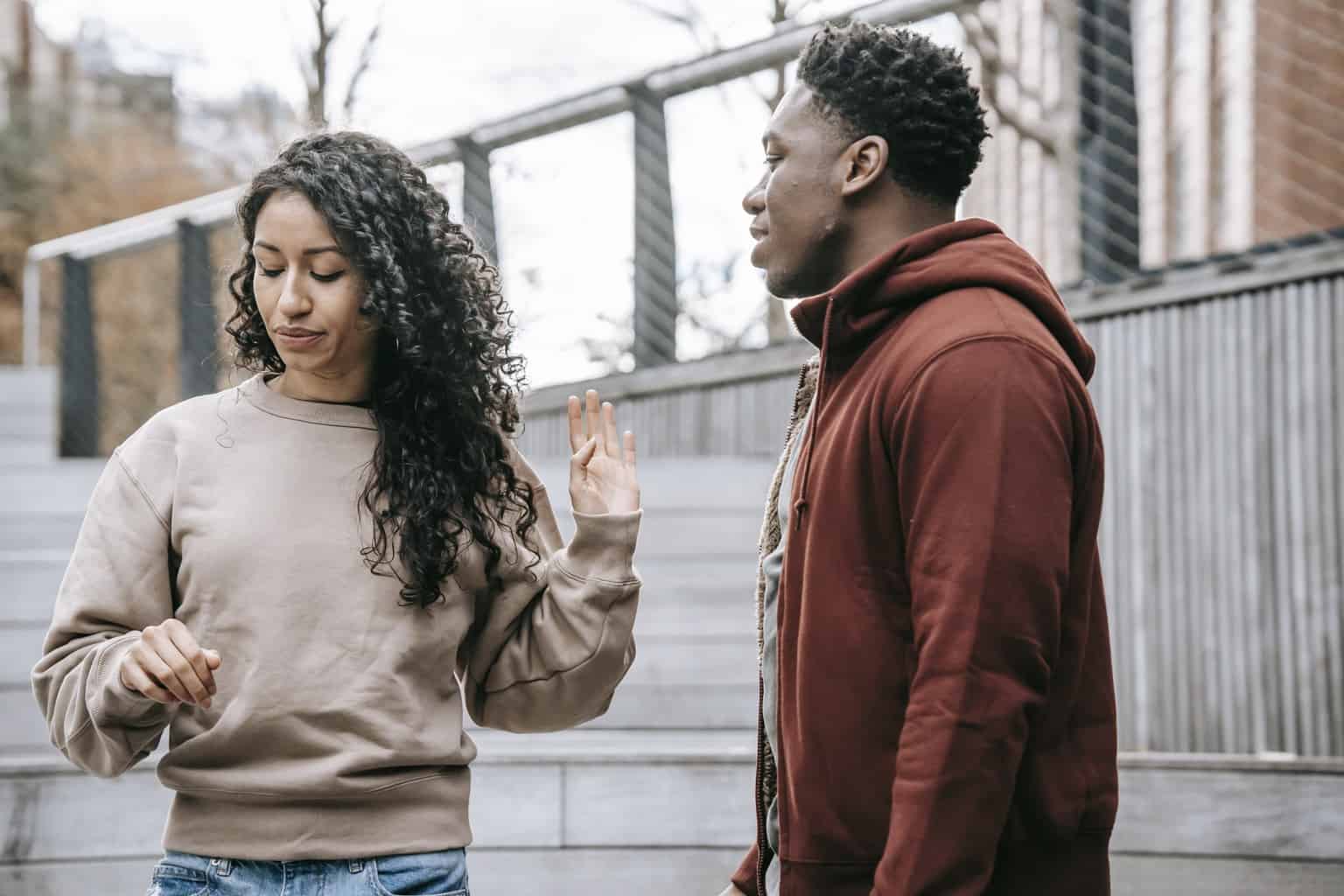 Upset ethnic girlfriend with raised hand and concerned African American boyfriend breaking up on street against metal grid on background