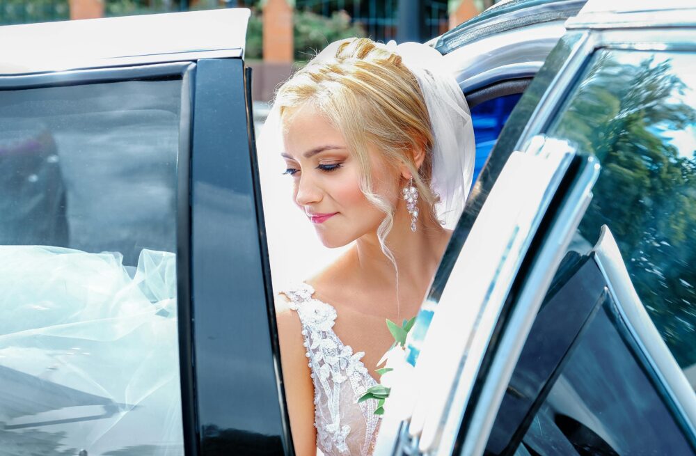 Charming young bride in white dress and veil sitting in black modern car on sunny summer day 