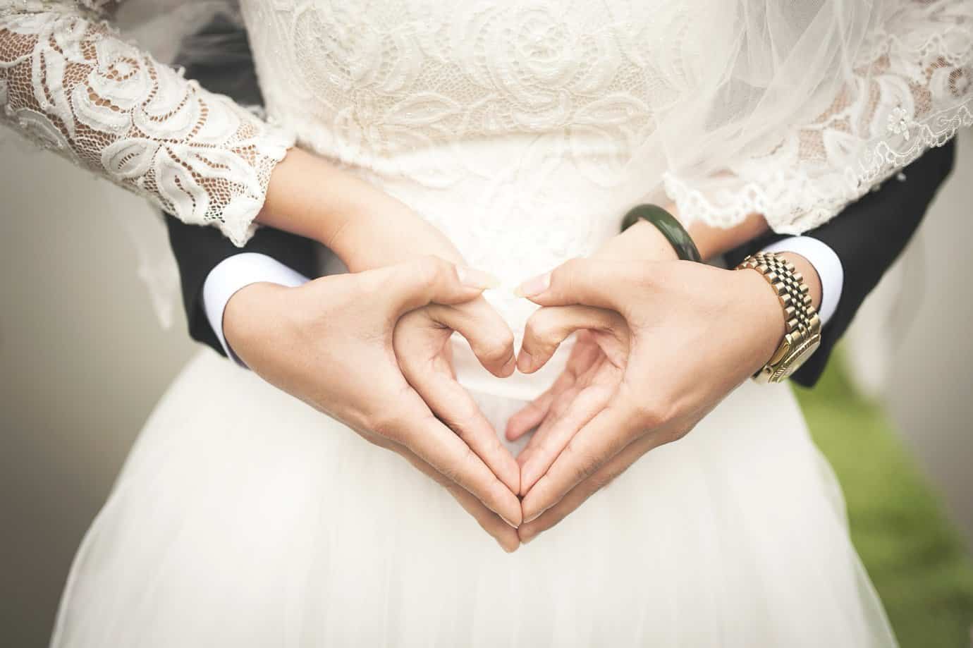 A couple in a wedding dress making a heart with their hands Description automatically generated