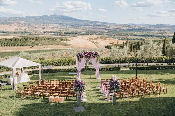 arch, decorated with trunks and flowers, stands in the wedding ceremony area on the hillside in Tuscany, as are the chairs for guests