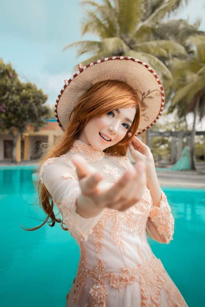 Free Beautiful young Asian bride with dyed red hair wearing lace white dress standing on poolside in tropical resort while outstretching hand and looking at camera Stock Photo